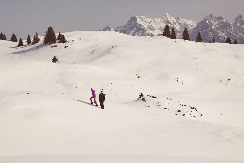 Two people walk across a snowy field with evergreen trees and snowy mountains in the background on a bright day in winter.