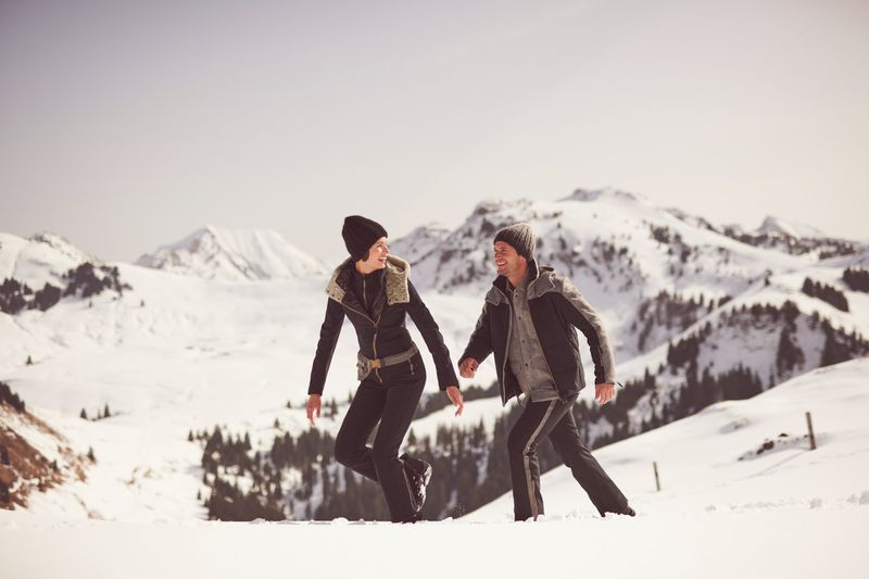 A couple walks in a snowy, mountainous landscape. They are wearing warm winter clothes and walking through a snowy field with mountains in the background.