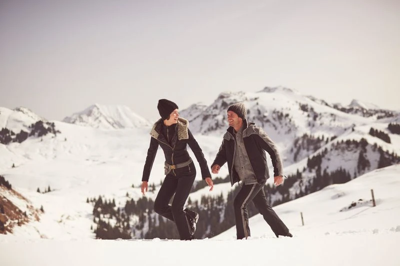 A couple walks in a snowy, mountainous landscape. They are wearing warm winter clothes and walking through a snowy field with mountains in the background.