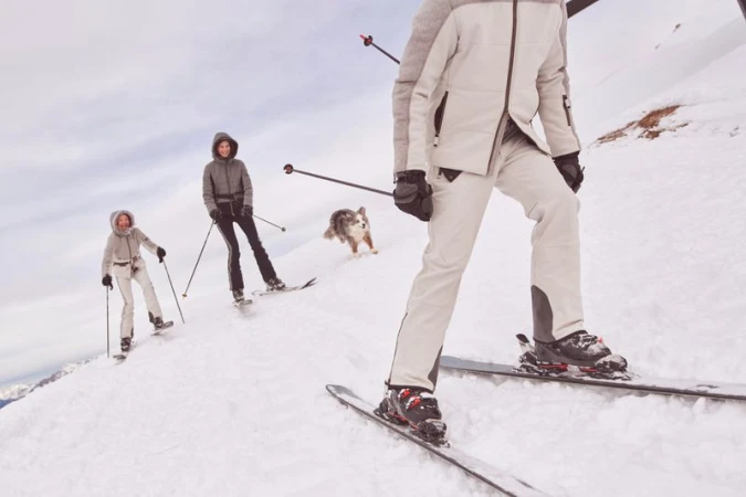 Three people skiing down a snowy mountain slope, with a dog running behind them under a bright sky in an outdoor environment.
