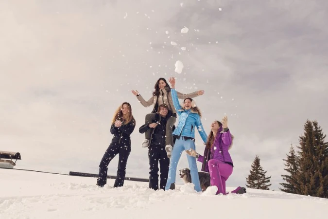 Five people are enjoying a winter day playing in the snow, with one man carrying a woman on his shoulders in an outdoor landscape.