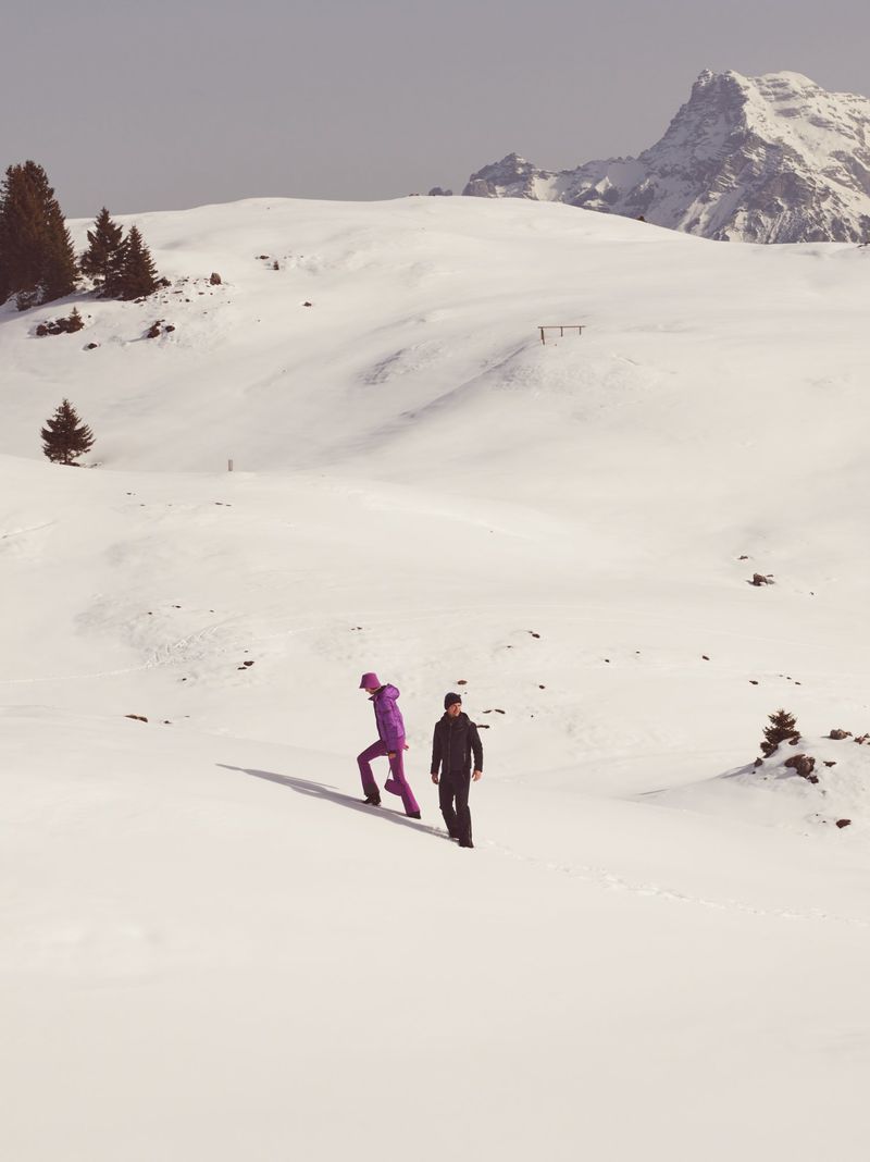 Two people walking uphill on a vast snowfield beneath distant snowy mountain peaks under an overcast sky.