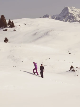 Two people walking uphill on a vast snowfield beneath distant snowy mountain peaks under an overcast sky.