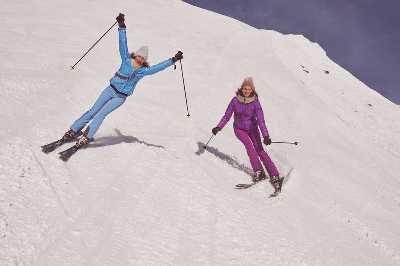 Two smiling women in winter ski gear enjoy skiing down a snowy mountain slope on a sunny day, showcasing a blend of fashion and winter sports.