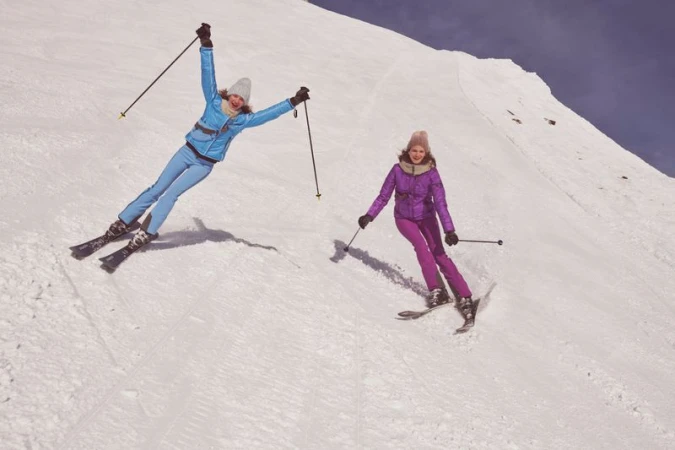 Two smiling women in winter ski gear enjoy skiing down a snowy mountain slope on a sunny day, showcasing a blend of fashion and winter sports.