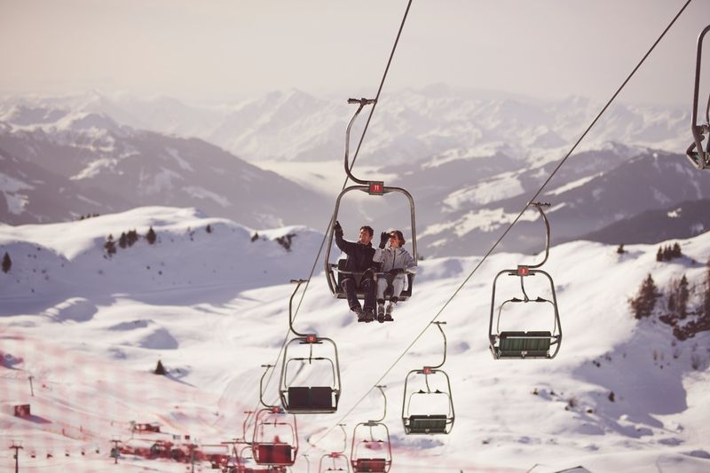 A couple rides a ski lift in a snowy mountain landscape on a sunny day. The ski lift is surrounded by snow and mountains.