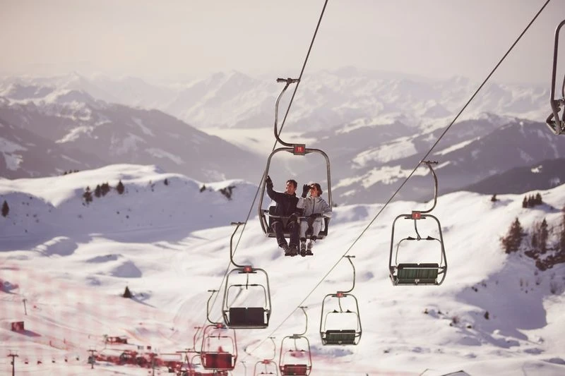 A couple rides a ski lift in a snowy mountain landscape on a sunny day. The ski lift is surrounded by snow and mountains.