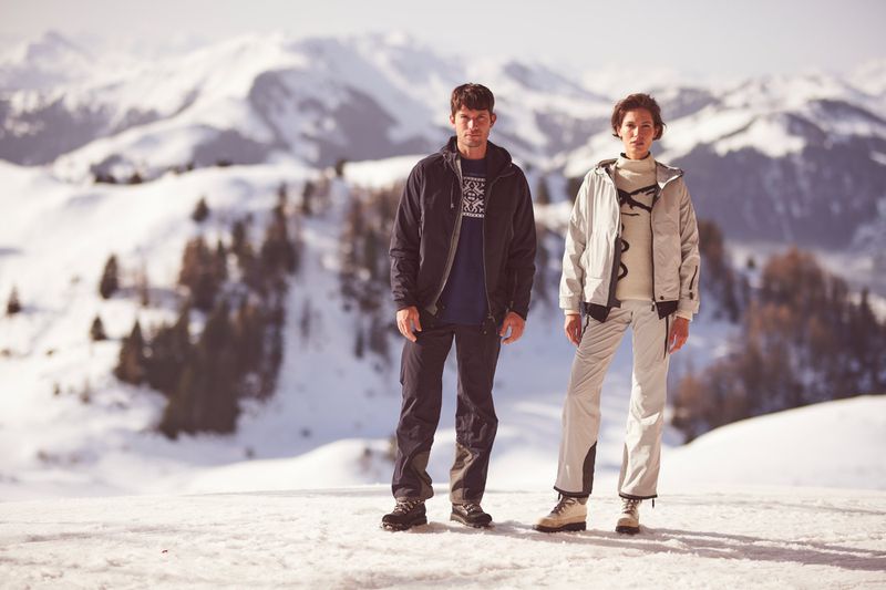 Two people pose on a snowy mountain wearing ski apparel in a medium shot, showcasing winter fashion and a snowy mountain backdrop.