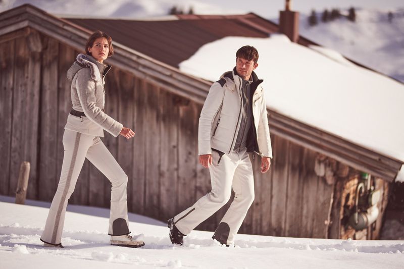 A couple in winter apparel walk in the snow near a wooden cabin, showcasing fashion and outdoor lifestyle in a scenic snowy mountain environment.