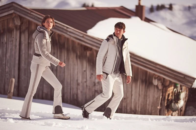 A couple in winter apparel walk in the snow near a wooden cabin, showcasing fashion and outdoor lifestyle in a scenic snowy mountain environment.