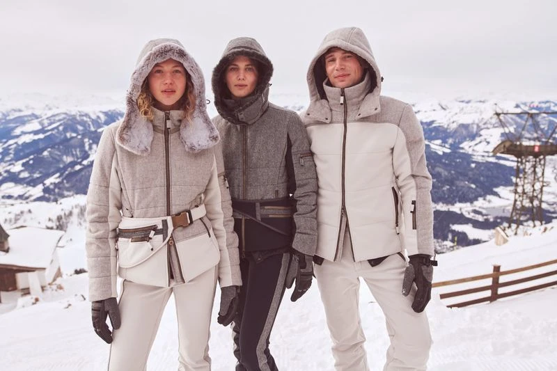 Three models in hooded winter ski jackets and snow pants pose in a snowy mountain landscape with a ski lift, showcasing winter fashion.