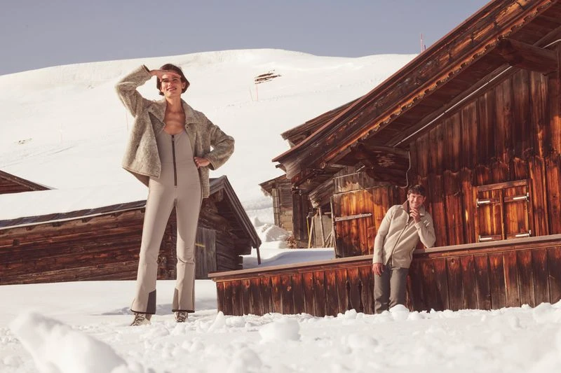 A woman in a ski suit stands on a snowy hill with an older man leaning against a wooden building on a sunny day.