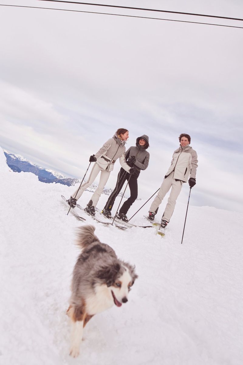 Three women skiing down a snowy slope on a cloudy day with a dog running in front of them toward the camera. 