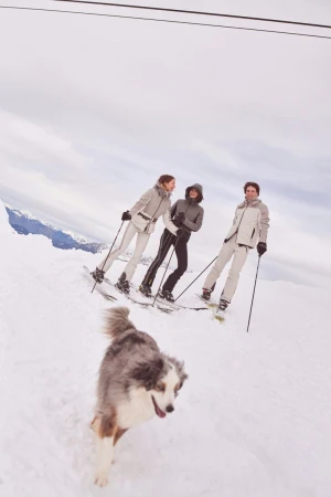 Three women skiing down a snowy slope on a cloudy day with a dog running in front of them toward the camera. 