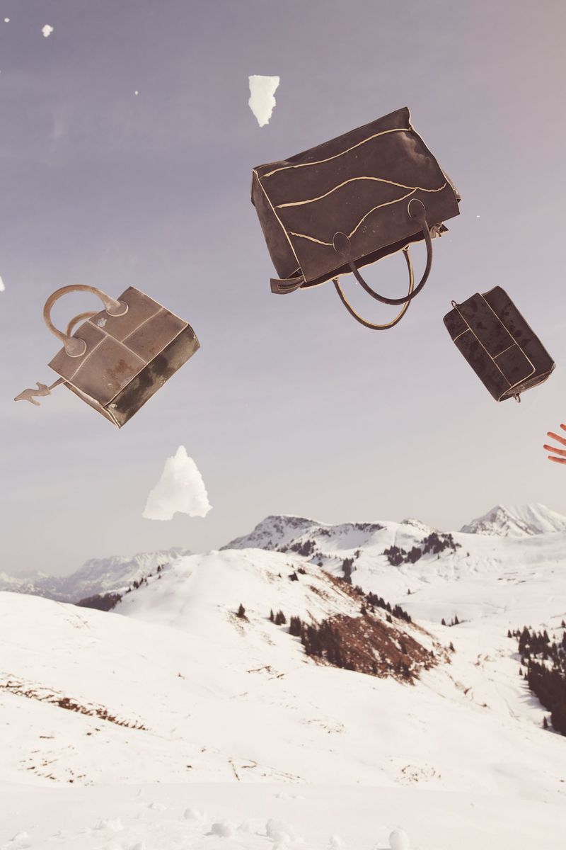 Whimsical fashion image: Three brown handbags in mid-air against snow-covered mountains and sky, suggesting a playful, gravity-defying scene.
