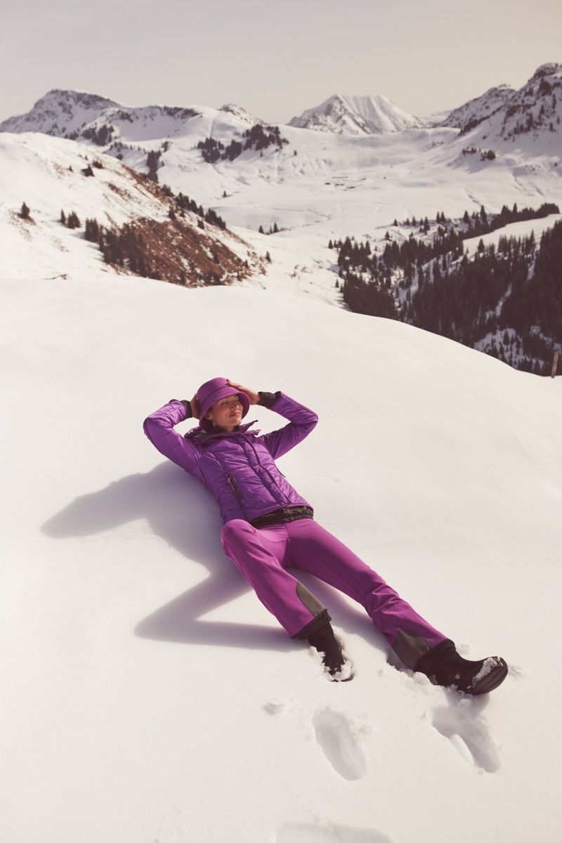 Woman in purple ski suit lying on snow in a sunny mountain landscape. Relaxing on slope with snow-covered mountain range in the background.