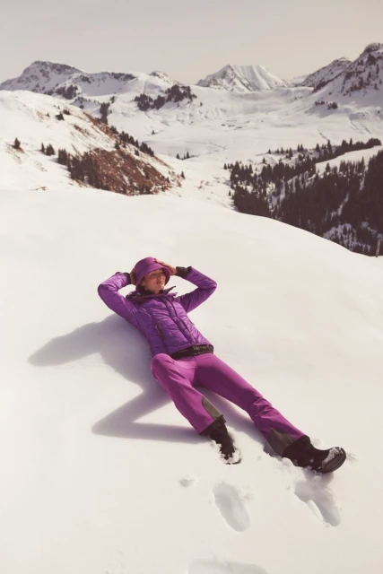 Woman in purple ski suit lying on snow in a sunny mountain landscape. Relaxing on slope with snow-covered mountain range in the background.