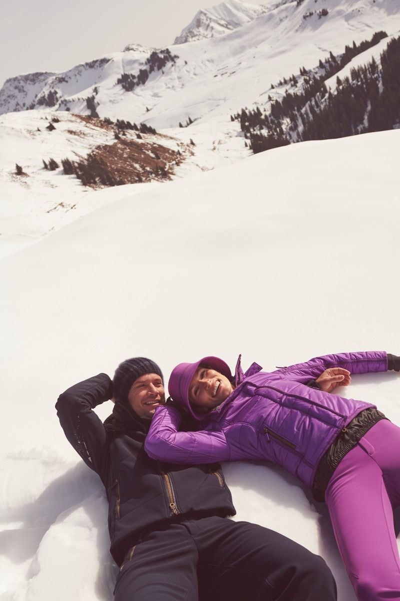 A couple lies in the snow with mountains in the background, wearing winter clothing, enjoying a sunny day in a winter wonderland.