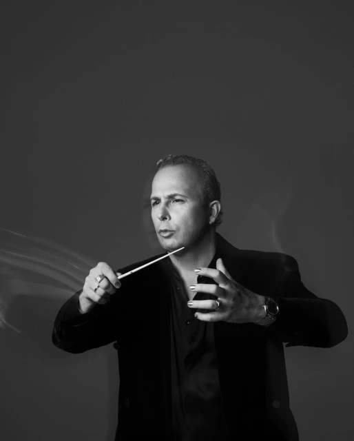 Monochrome portrait of a conductor in a black velvet jacket holding a baton with hands gesturing in front of a neutral background.