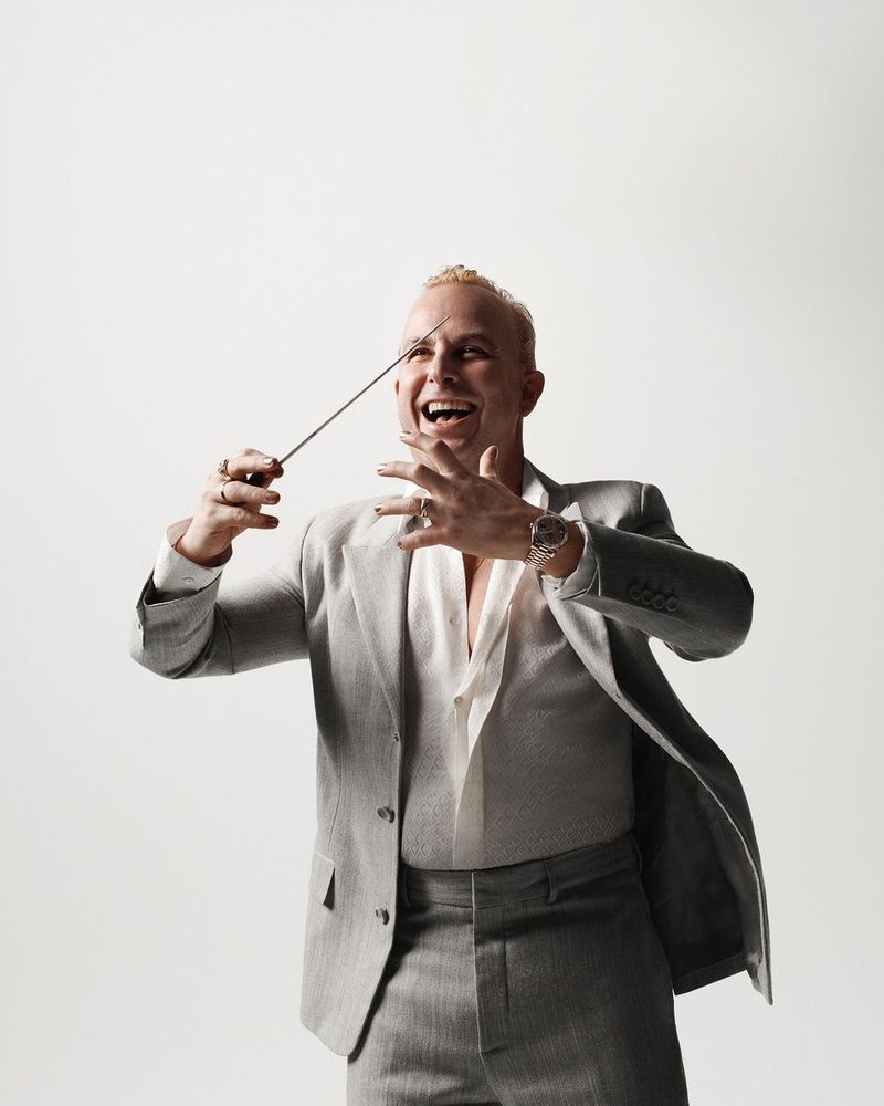 Man in grey suit smiles, conducting with baton against a bright background in studio lighting, showcasing a blend of formal wear and energetic expression.