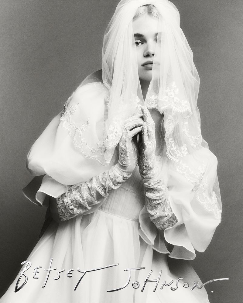 Monochrome fashion portrait of a fair-skinned model wearing a white lace-trimmed bridal gown with veil, set against a grey background.
