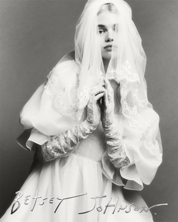 Monochrome fashion portrait of a fair-skinned model wearing a white lace-trimmed bridal gown with veil, set against a grey background.