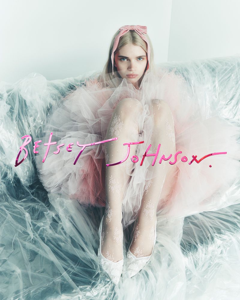 A young woman sits on a plastic-covered couch wearing a pink tulle dress, lace shoes, and a pink bow headband. She looks at the camera.