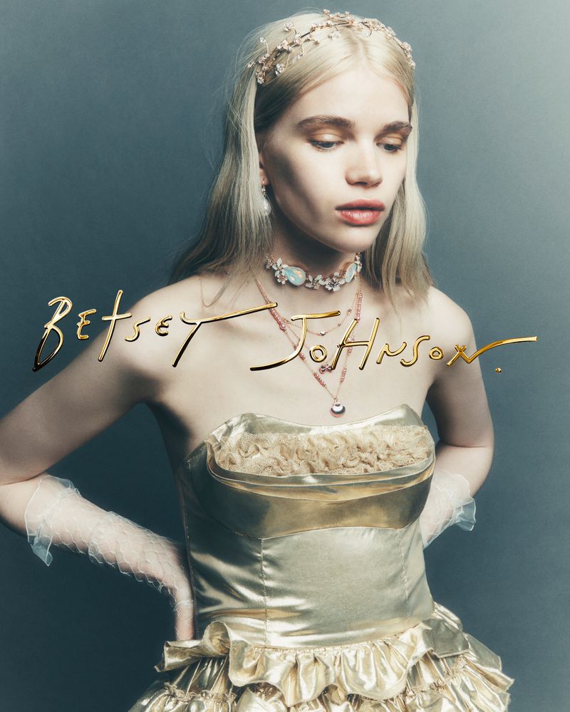 An adult female fashion model poses in a gold dress and jewelry by Betsey Johnson against a grey background in a studio shot.