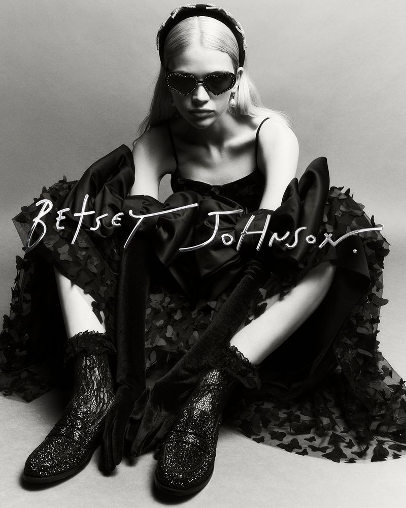 Monochrome studio shot of a young adult woman in a black dress, heart-shaped sunglasses, and platform boots, sitting and posing.