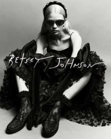 Monochrome studio shot of a young adult woman in a black dress, heart-shaped sunglasses, and platform boots, sitting and posing.