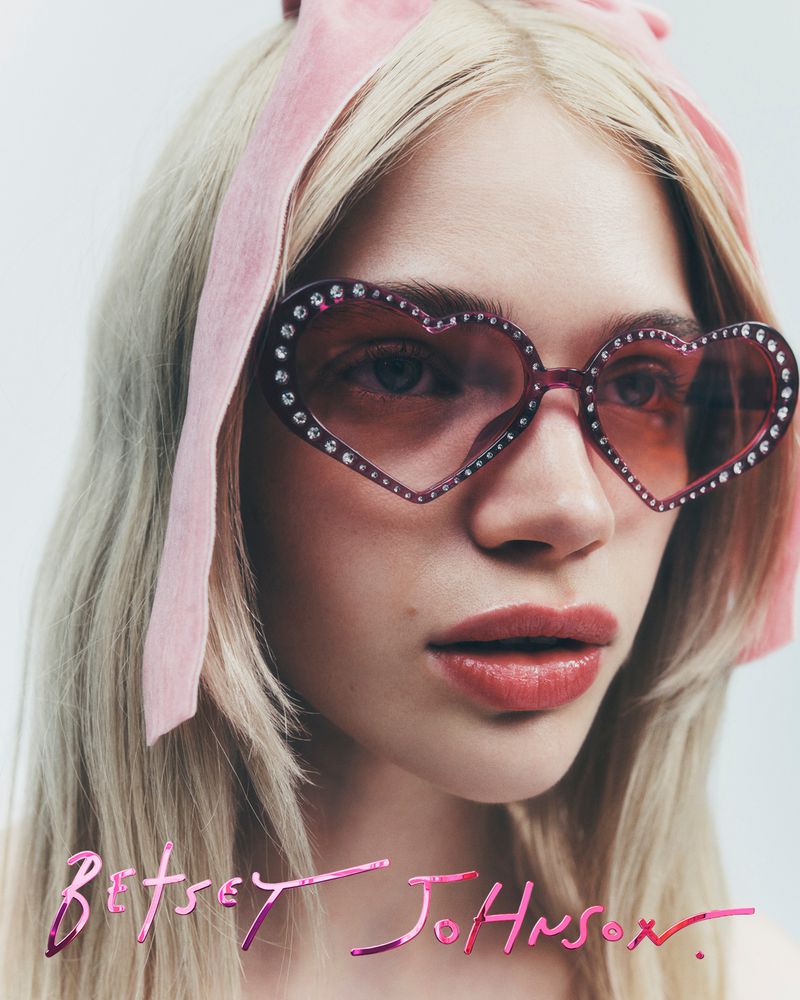 Close-up of a blonde woman wearing heart-shaped sunglasses with pink frames and rhinestones, complemented by a pink velvet hair bow.