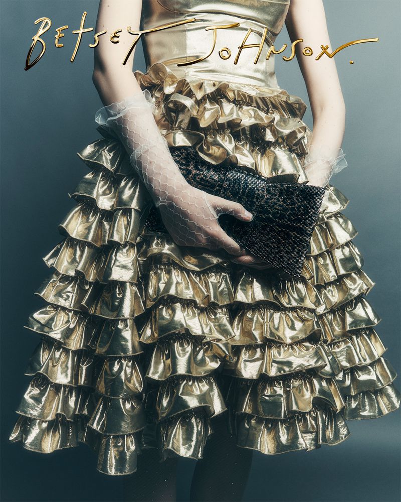 Woman in a shiny gold dress with ruffles holds a textured clutch, accessorized with sheer gloves, against a grey background, fashion photography.