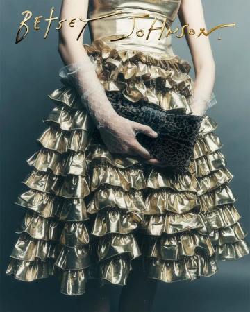 Woman in a shiny gold dress with ruffles holds a textured clutch, accessorized with sheer gloves, against a grey background, fashion photography.