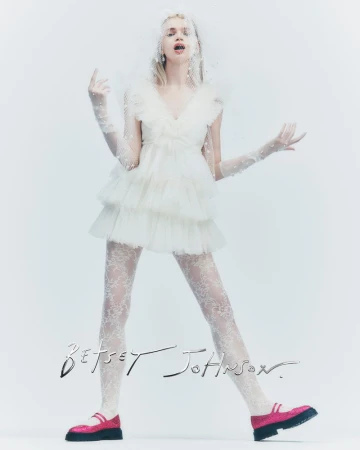 A woman poses in a white dress, lace leggings, and pink shoes against a white background in a fashion editorial shot.