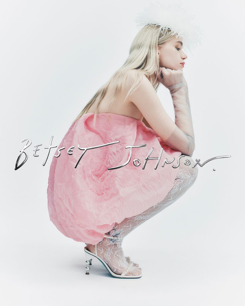 Woman in pink dress and sheer heels, crouching in a studio setting. She has blonde hair and is wearing sheer gloves.