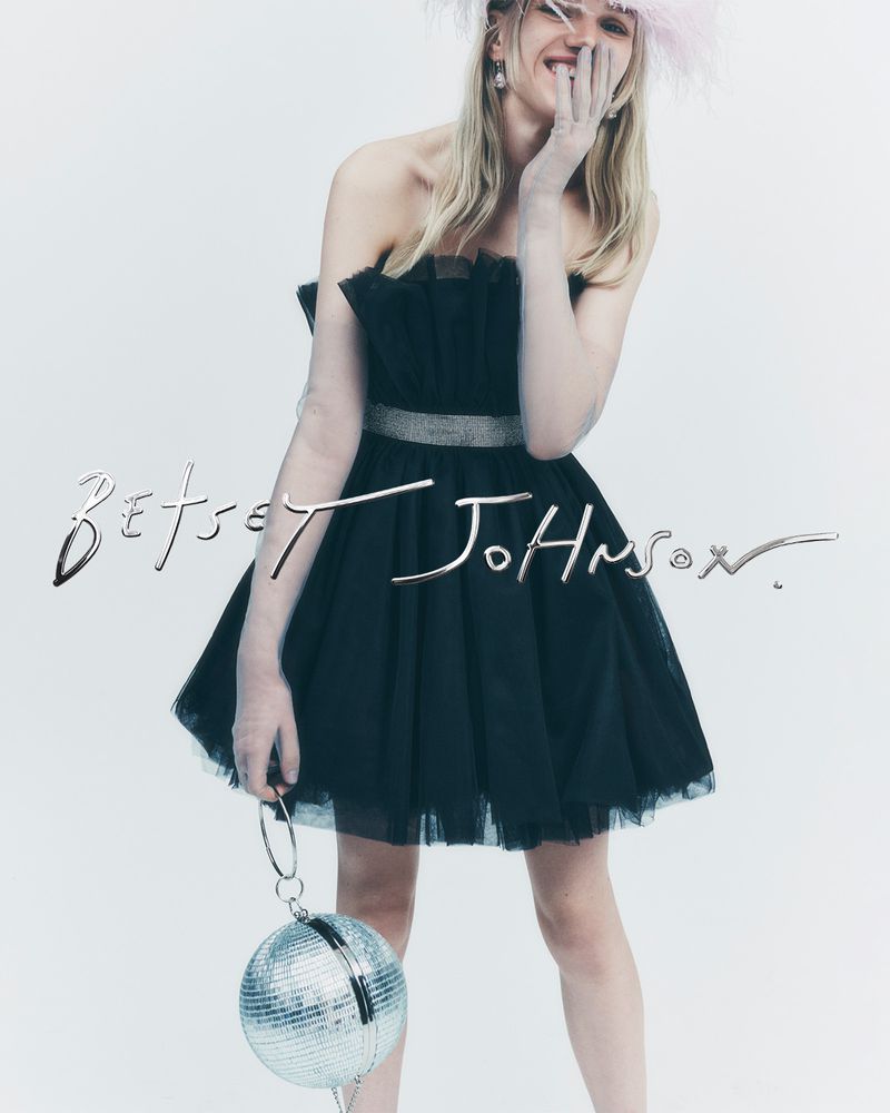 A joyful woman in a black tulle dress and feather hat poses against a white background holding a disco ball purse.