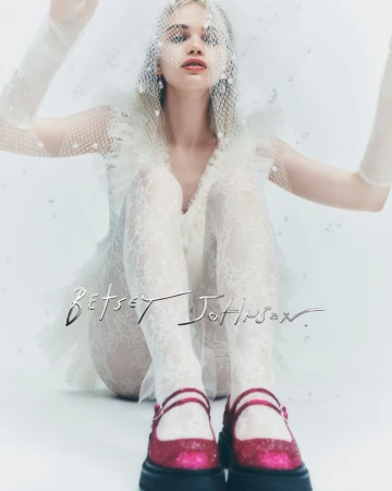 A young woman in a white dress and veil poses in a bright, high-key studio wearing glittery pink Mary Jane shoes and white lace tights.