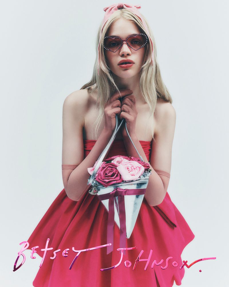 A model showcases a Betsey Johnson design: heart-shaped sunglasses, a pink dress, and a cone-shaped purse filled with pink roses on a white background.