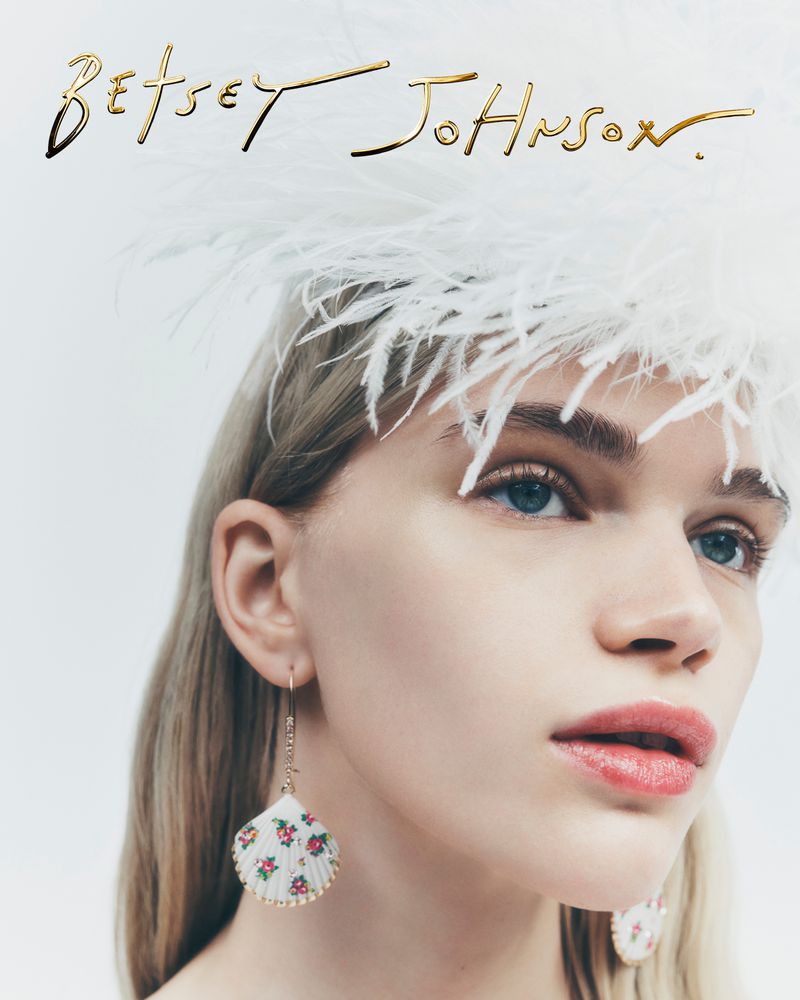 A close-up shot of a fair-skinned adult female wearing a feathered headpiece and shell-shaped earrings, complemented by 'Betsey Johnson' branding.