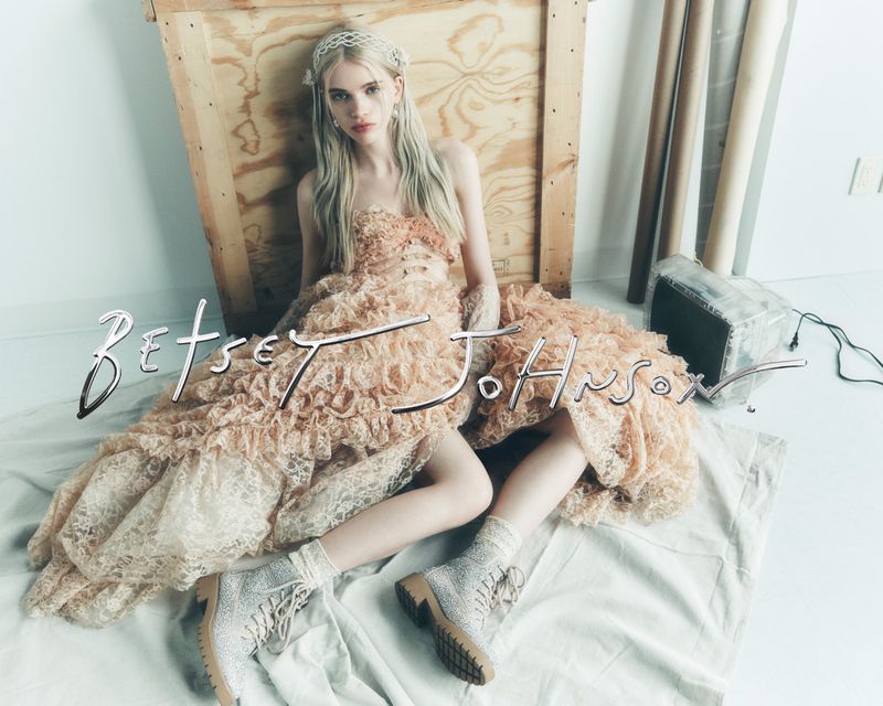A young woman poses in a peach tulle dress and sparkling boots, sitting in a studio with wooden crates and paper rolls in the background.