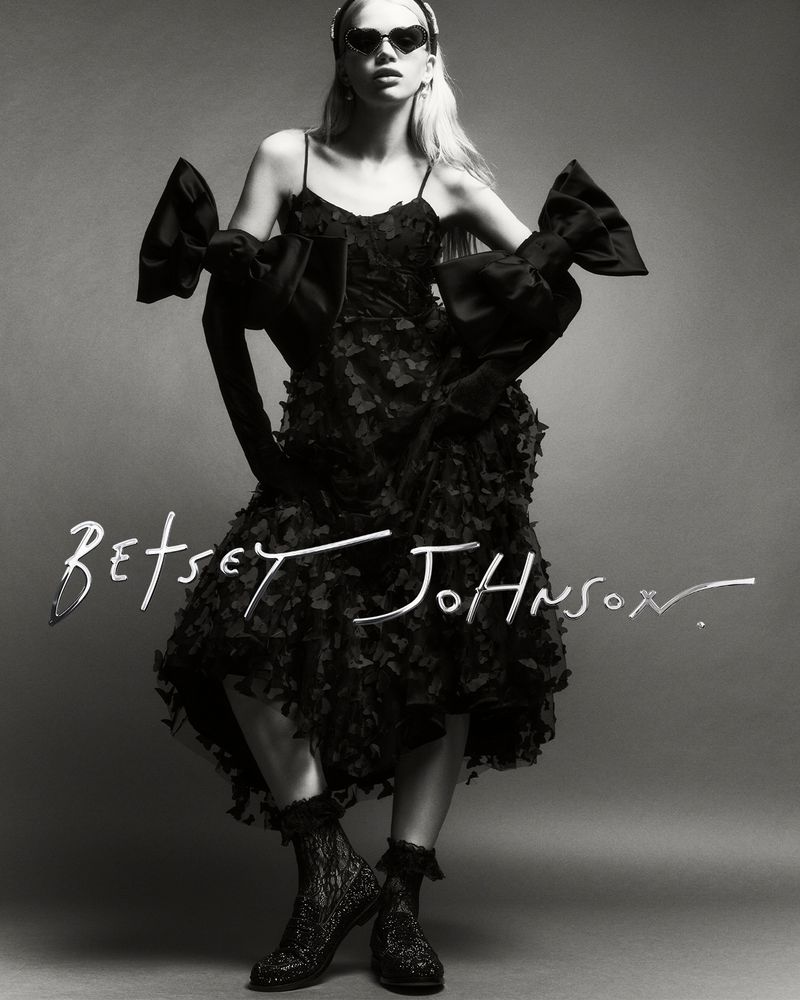 Black and white fashion shot of a woman in a Betsey Johnson dress with large bows and heart-shaped sunglasses, posing in a studio.