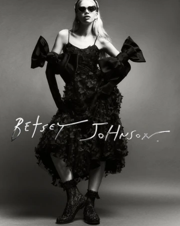 Black and white fashion shot of a woman in a Betsey Johnson dress with large bows and heart-shaped sunglasses, posing in a studio.
