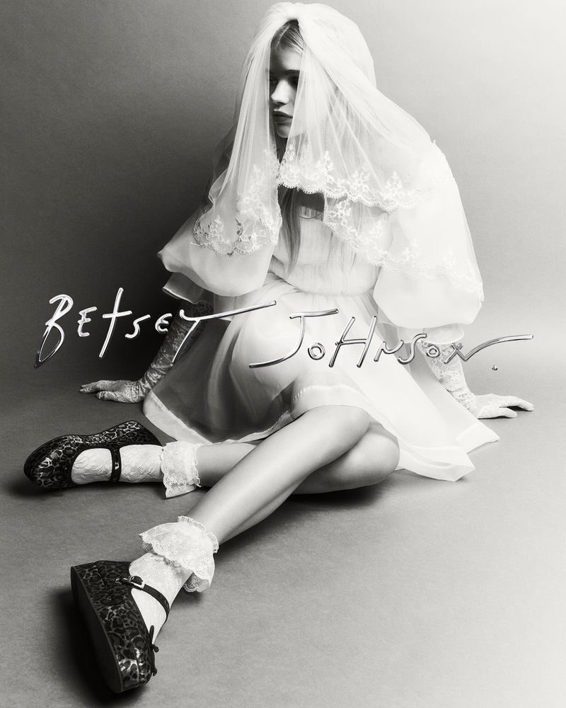 Woman in a bridal-style dress with a veil and Mary Jane platform shoes, posing in front of a plain grey backdrop, and Betsey Johnson text overlay.