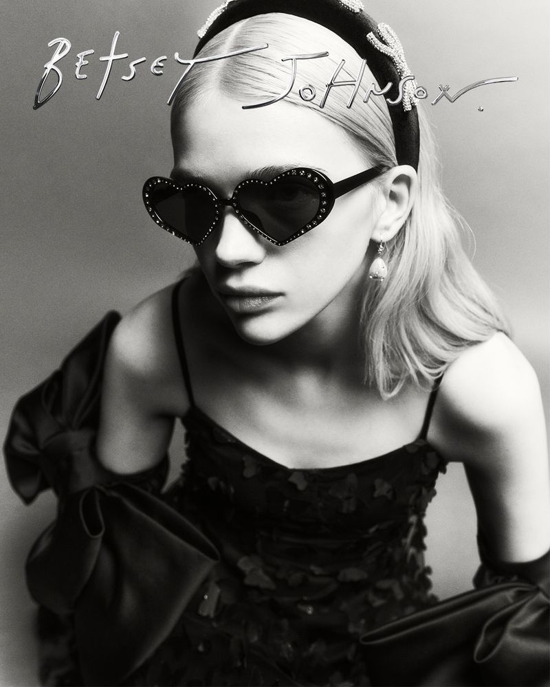 Monochrome portrait of a blonde woman wearing heart-shaped sunglasses, a black dress, and a headband, with 'Betsey Johnson' text overlay.
