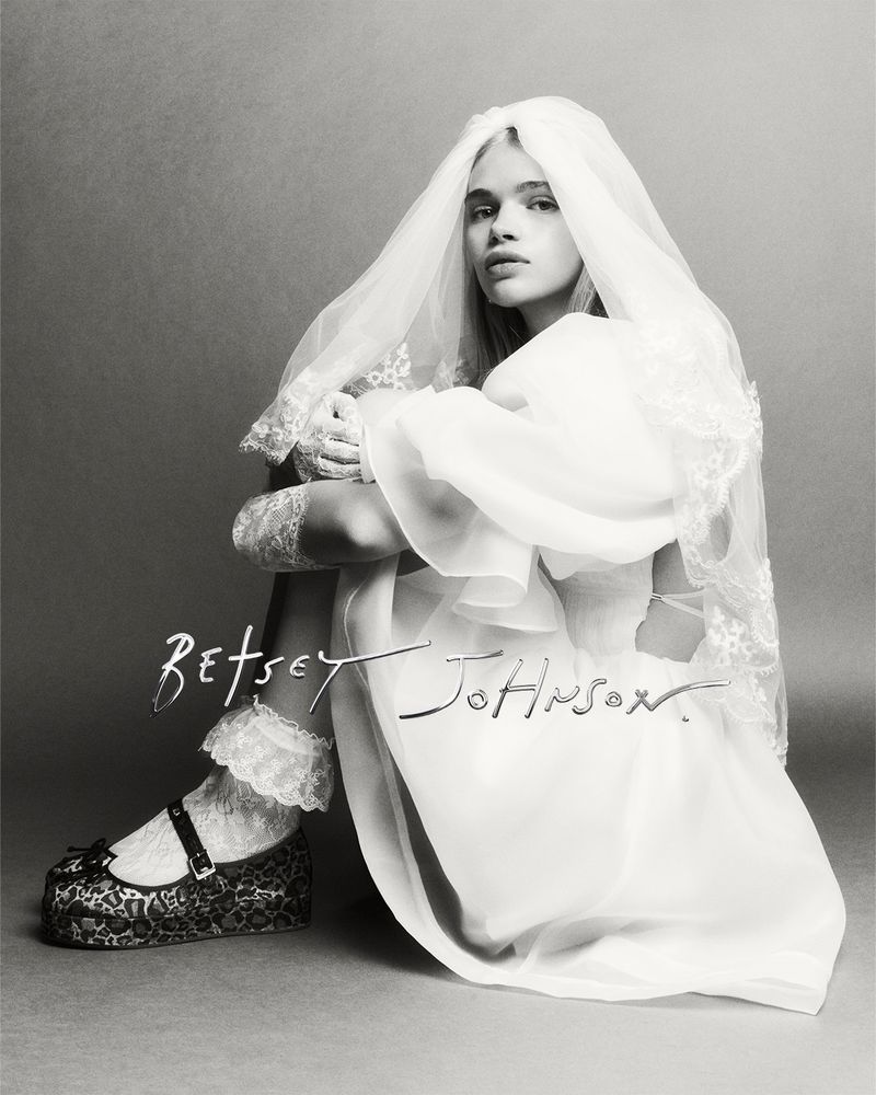 Monochrome fashion shot of a young woman in a sheer white bridal dress and veil, sitting on the floor in a studio setting.