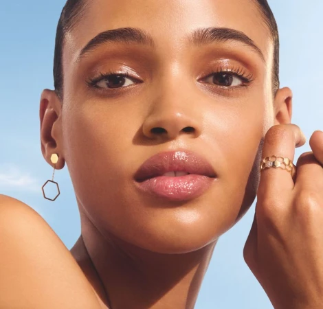 Close-up beauty shot of an adult woman with gold hexagon earrings and honeycomb ring against a pale blue sky backdrop.