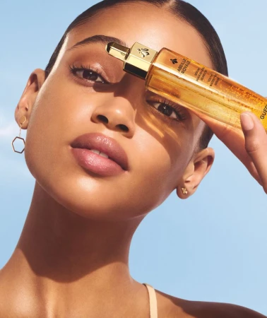 Close-up of a woman with tan skin and dark hair posing with a bottle of Guerlain Abeille Royale skincare serum against a light blue background.