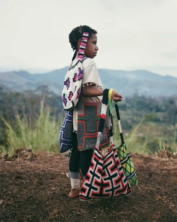 Child in Papua New Guinea stands on a hillside wearing traditional patterned bags, facing left, with a blurred mountain background.