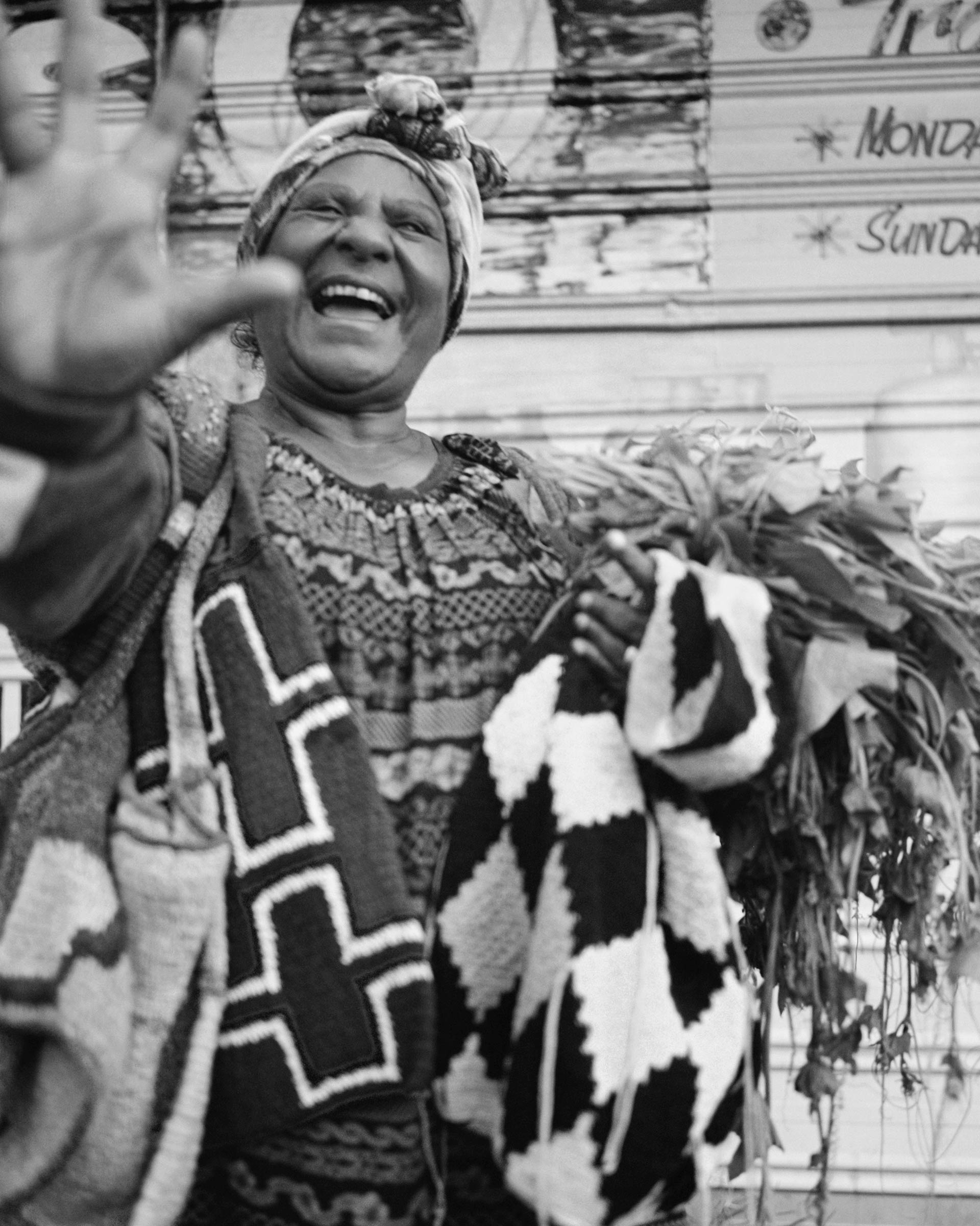 Black and white portrait of a smiling older woman wearing a patterned dress and headscarf, holding foliage and a patterned bag.