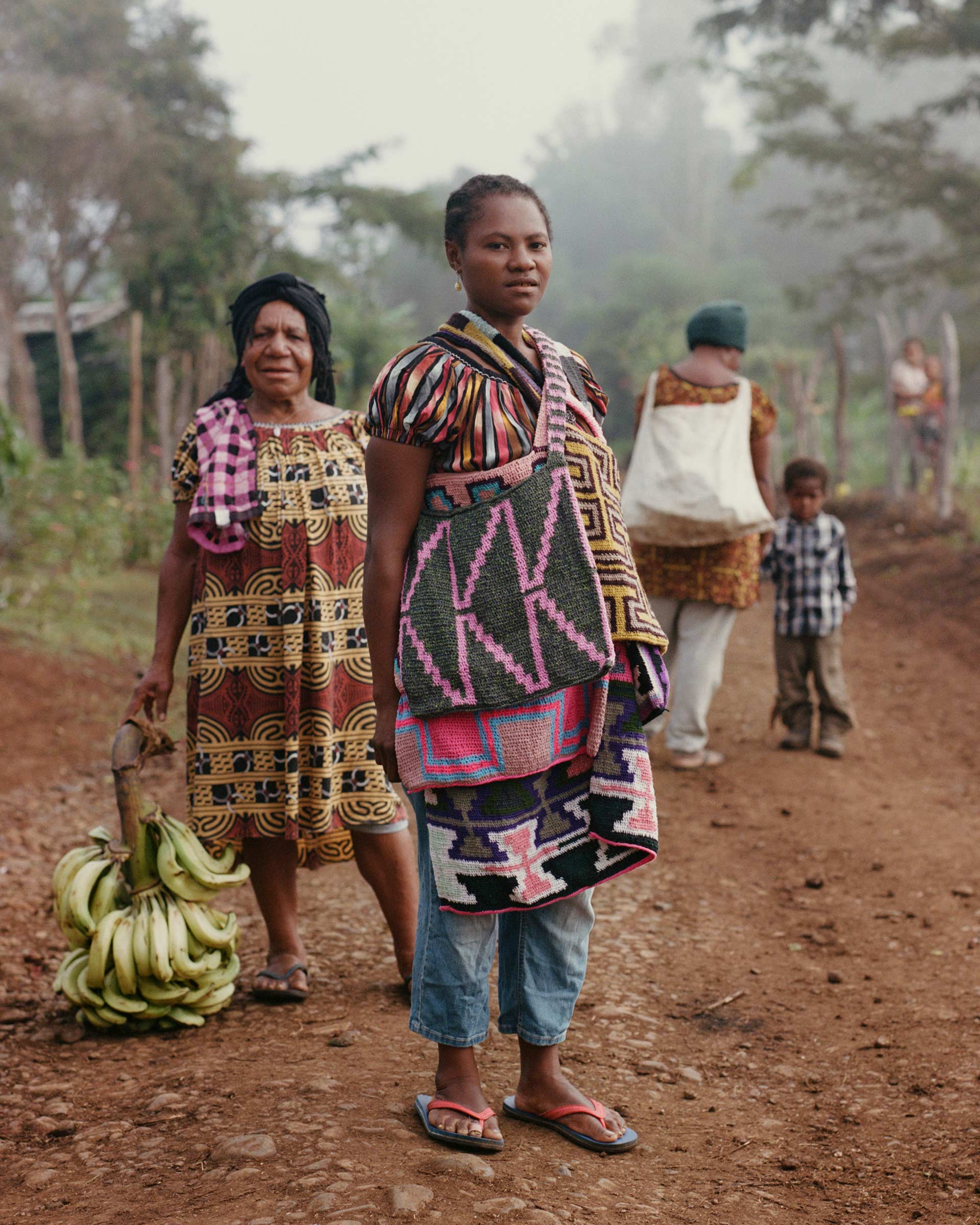 Three women and a child stand on a dirt road near lush greenery; an adult woman carries bananas and other colorful textile bags.